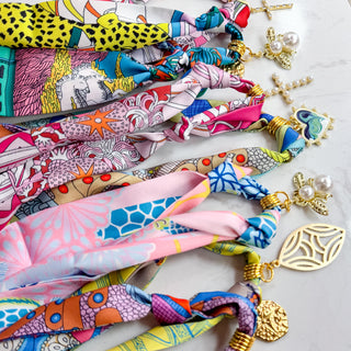 Assorted colorful scarf necklaces with knotted fabric and bold gold-tone charms including crosses, pearls, and medallions