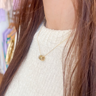 Gold necklace with two pendants worn by a person with long brown hair, against a blurred background.