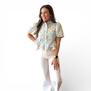 Light blue floral blouse with tan botanical print, puff sleeves, button front, and peplum hem styled with white pants on a smiling model against a clean background