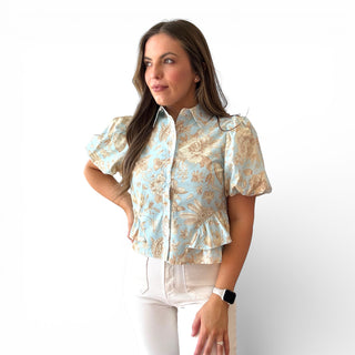 Light blue floral blouse with tan botanical print, puff sleeves, button front, and peplum hem styled with white pants on a model against a clean background