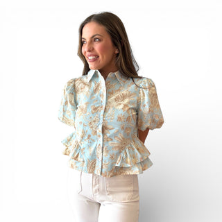 Light blue floral blouse with tan botanical print, puff sleeves, structured collar, and peplum hem styled with white pants on a smiling model against a clean background