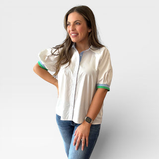 White button front blouse with contrast blue placket and collar, puff sleeves with green trim, styled with medium wash jeans on a smiling model against a clean background