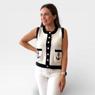 Cream sleeveless knit vest with black trim, gold button front, and anchor detail pockets styled with white pants on a smiling model against a clean background