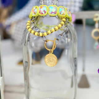 Kitzi Victoria mixed metal coin bracelet with silver-tone chain, gold toggle and coin charm, styled in a colorful bracelet stack on glass display