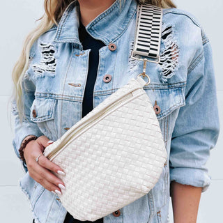 Woman wearing a distressed denim jacket and holding a cream woven sling bag with a wide black and white striped adjustable strap.