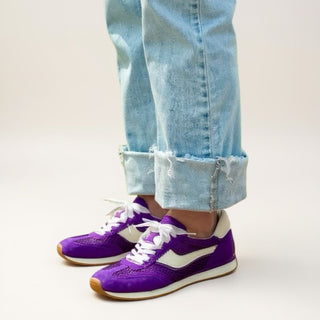 Purple sneakers with white laces worn with light blue jeans on a plain background