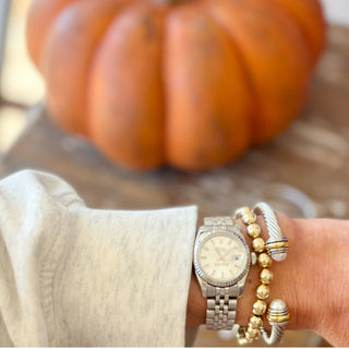 Hand wearing a watch and silver bracelet with pearl detailing with a pumpkin in the background