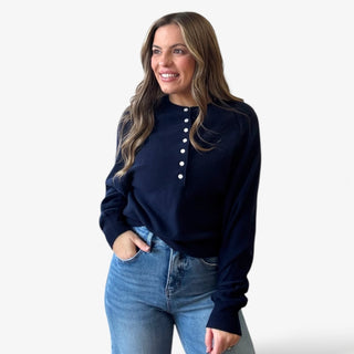 Woman wearing a navy blue long-sleeve sweater top with white buttons and light blue jeans on a white background
