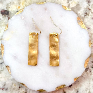 Hammered gold bar drop earrings with a brushed finish and fishhook backs, displayed on a white stone slab with gold edging.