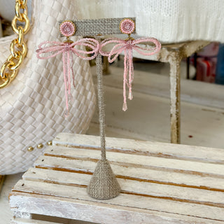 Blush pink beaded bow drop earrings with dangling tails displayed on neutral jewelry stand