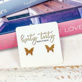 Gold butterfly stud earrings displayed on a white Hoity Toity Boutique card, styled in front of stacked coffee table books with pink and blue covers.