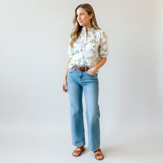 White and green botanical print blouse with button front, short puff sleeves, and split neckline styled with light wash straight leg jeans, brown belt, and flat sandals on a model against a clean background