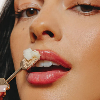 Close-up of a woman's face with glossy pink lips as she holds a small poppy & pout lip care spatula with lip mask near her mouth.