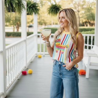 Woman in a queen of sparkles pinata colorful striped tank top and jeans holding a drink on a porch.
