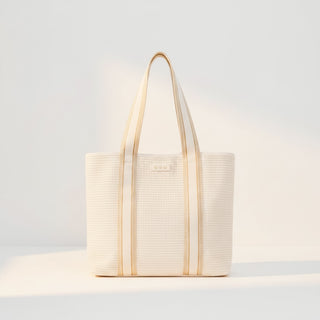 Ivory raffia tote bag with neutral striped straps and woven texture shown in a minimalist studio setting.