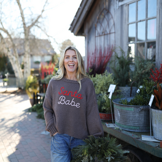 Woman wearing a sweater with 'Santa Babe' text in an outdoor setting with plants and a building.