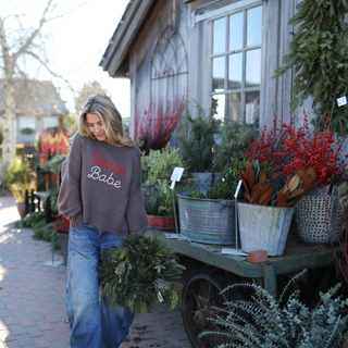 Woman wearing a 'Santa Babe' sweater standing next to a cart of plants in an outdoor setting.