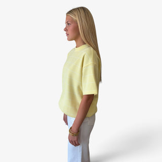 A woman in profile wearing a pale yellow short-sleeve scuba knit top with thin white horizontal stripes and white pants, from a angled view