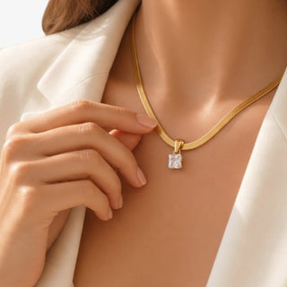 Gold pendant necklace with square clear crystal charm and sleek flat snake chain, modeled with a cream blazer, close-up of delicate layering jewelry.