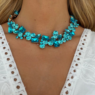 Teal blue crystal statement collar necklace with mixed gemstone shapes on gold setting, styled on model with white eyelet top, front view.