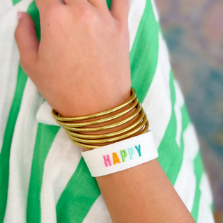 Tie Happy white fabric bracelet embroidered with colorful HAPPY lettering, styled with stacked gold bangles on wrist