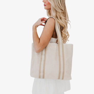 Cream raffia tote bag with beige striped shoulder straps worn over shoulder by blonde model side view.