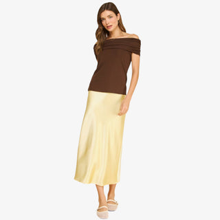 Woman wearing a butter yellow satin midi slip skirt styled with a brown top and neutral flats on a white background