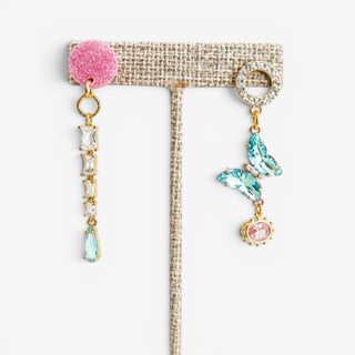 Blue butterfly mismatched statement earrings with pink glitter studs, clear crystals and aqua teardrop accents displayed on an earring stand.