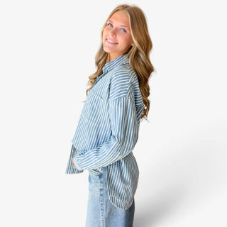 Women's blue striped button down shirt featuring contrasting stripe placement, dropped shoulders, button cuffs, and a curved hem, side view with jeans.