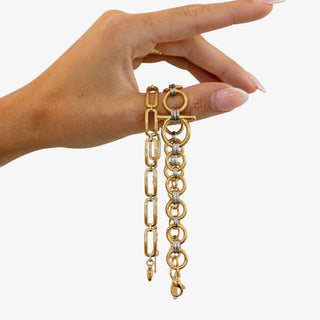 Close-up of a hand holding two delicate gold bracelets; one has elongated links and the other has round links with silver bands against a background.