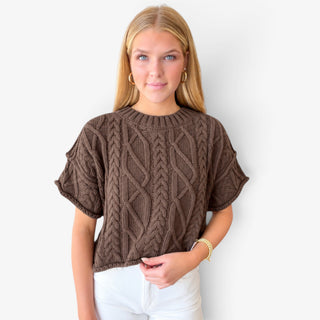 Brown cable knit sweater with short sleeves and textured braided knit pattern, front view on model with white pants.
