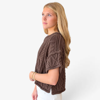 Brown cable knit sweater with relaxed short sleeves and textured cable design, side view on model with white pants.