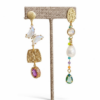 Gold mismatched statement earrings with an iridescent butterfly, pearl, colorful beads and pink and green crystal drops shown on an earring stand.