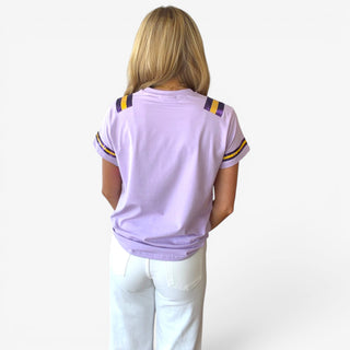 Back view of lavender short sleeve game day tee with purple and gold striped sleeve and shoulder details styled with white jeans