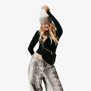 Woman holding a Pretty Simple Suede Westlyn Woven Bum Bag in rich chocolate brown with gold zipper detail, styled with a black one-shoulder top and printed pants.
