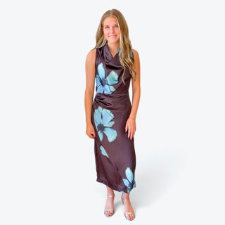 Women's dark brown floral midi dress with oversized blue flowers, a draped cowl neckline and sleeveless design, styled with silver heels, full front view.