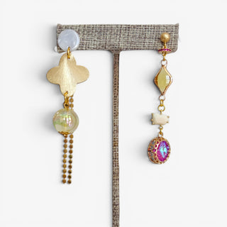 Gold and pink mismatched statement earrings with clover, pearl, crystal and chain details displayed on an earring stand.