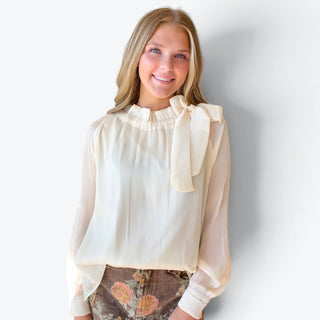 Women's ivory ruffle neck blouse with sheer long sleeves and statement shoulder bow, tucked into a brown floral skirt, front view.