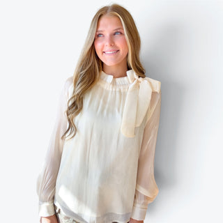 Ivory long sleeve blouse featuring a ruffled high neckline, oversized shoulder bow and relaxed flowing fit, styled with a floral skirt, front view.