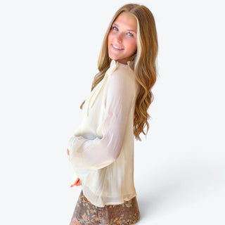 Ivory ruffle neck tie blouse with sheer balloon sleeves, gathered cuffs and a statement bow at the shoulder, modeled from the side.