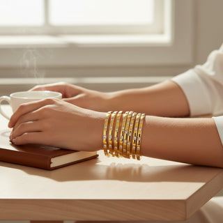 Person's hands with gold bracelets on a table holding a cup of coffee.