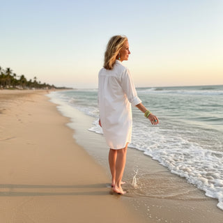 White Kaira Poplin Shirt Dress featuring a clean back design and relaxed fit, shown walking along the shoreline, back view.