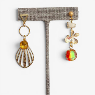 Orange and gold mismatched statement earrings with a shell-shaped drop, floral accent and iridescent crystal displayed on an earring stand.