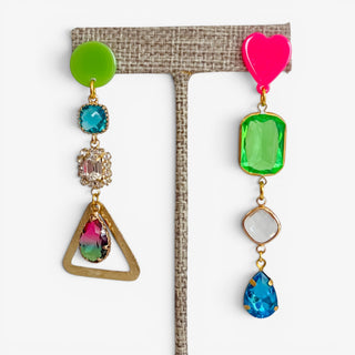 Pink and green mismatched statement earrings with colorful crystals, heart stud, geometric gold drop and blue teardrop displayed on an earring stand.