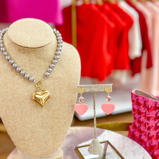 Pink and gold dangle heart earrings on a stand with a silver bead necklace and gold heart necklace
