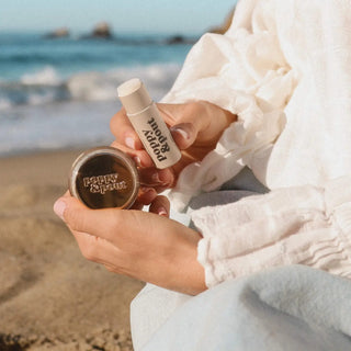 Woman holding Poppy & Pout Island Coconut lip balm and lip scrub at the beach for nourishing on-the-go lip care