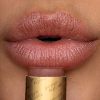 Close-up of a person's lips wearing  poppy & pout sheer lip tint, dusty pink lip product, with the gold-tipped tube of lip balm held at the bottom.