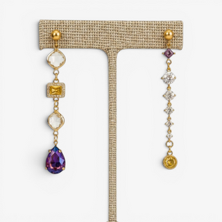 Purple and gold mismatched statement earrings with clear crystals, yellow accents and a purple teardrop gemstone displayed on an earring stand.