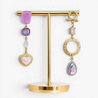 Purple and gold mismatched statement earrings with an iridescent heart, lavender crystals, hammered circle and rainbow teardrop displayed on a gold stand.