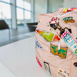 Close-up of beige quilted duffle bag with colorful sequin travel patches and embroidered strap detail by Queen of Sparkles 